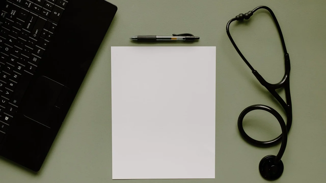 A blank clipboard beside a laptop and stethoscope