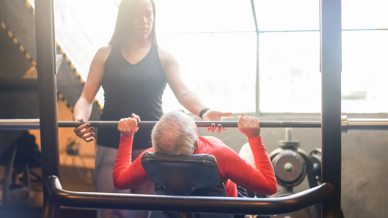 Older man using a chest-press machine beside a trainer.